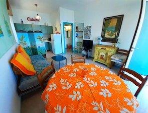 Ein Essbereich mit einem runden Tisch, der mit einer orangefarbenen Tischdecke bedeckt ist, einer Couch mit Kissen, einem Fernseher und einer Vitrine mit Dekorationsgegenständen.