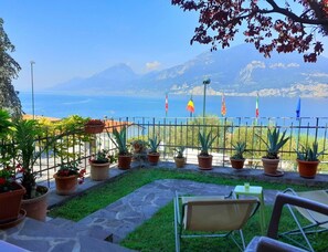 Eine Terrasse mit Kübelpflanzen, einem Tisch und Stühlen mit Blick auf die Küste, Berge und Flaggen.