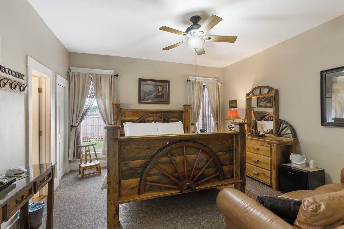 A bedroom with a wooden bed, a ceiling fan, a wooden dresser, a nightstand, and a window with curtains.
