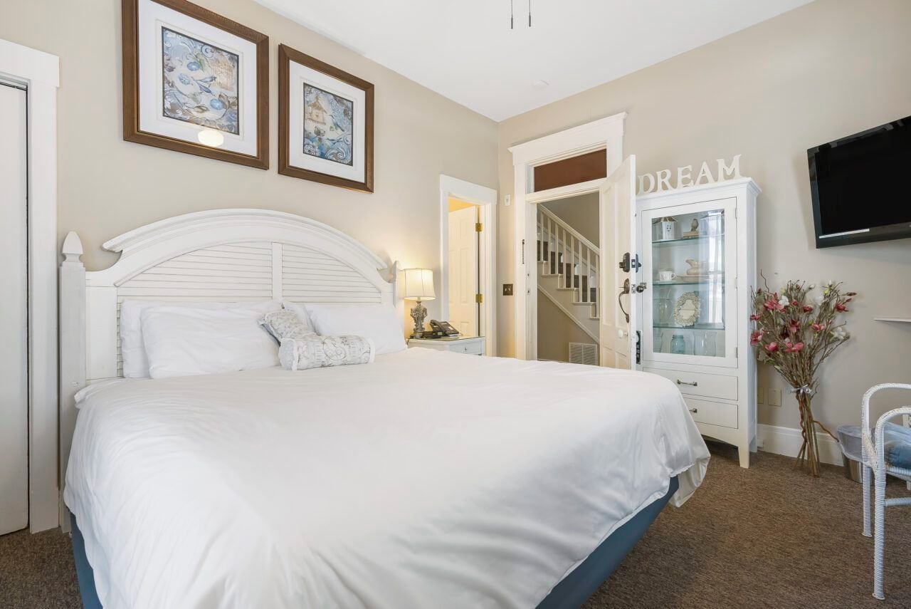 A bedroom with a large bed, a dresser, a television mounted on the wall, and a door leading to a staircase.
