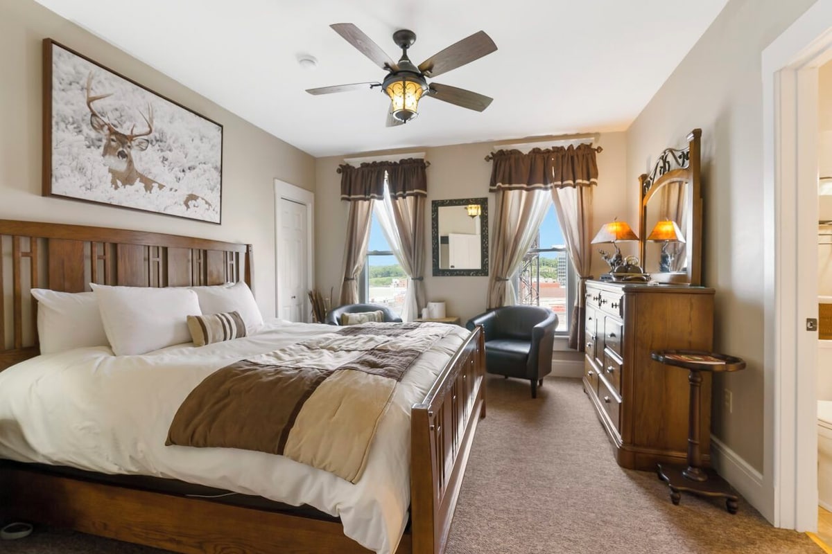 A hotel room with a large bed, a ceiling fan, a desk with a lamp, and a view of the outdoors.