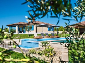 A poolside area with lounge chairs and umbrellas, a clear blue sky, and a building with a tiled roof. - Villa in Fazana With Swimming Pool (Fazana)