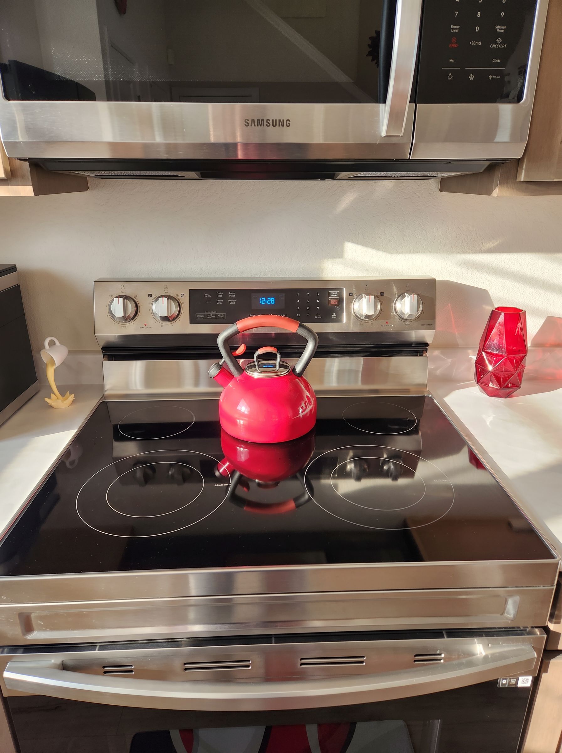 A stainless steel Samsung microwave above a stove with a red kettle on it.