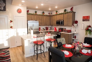 A modern kitchen with wooden cabinets, a stainless steel refrigerator, a microwave, and a red toaster. There are two bar stools with red cushions, a dining table with a red placemat, and a vase with red flowers on the counter. - 3-bedroom luxury townhome in charming Colorado Springs w/AC - Paradise Point (Colorado Springs)