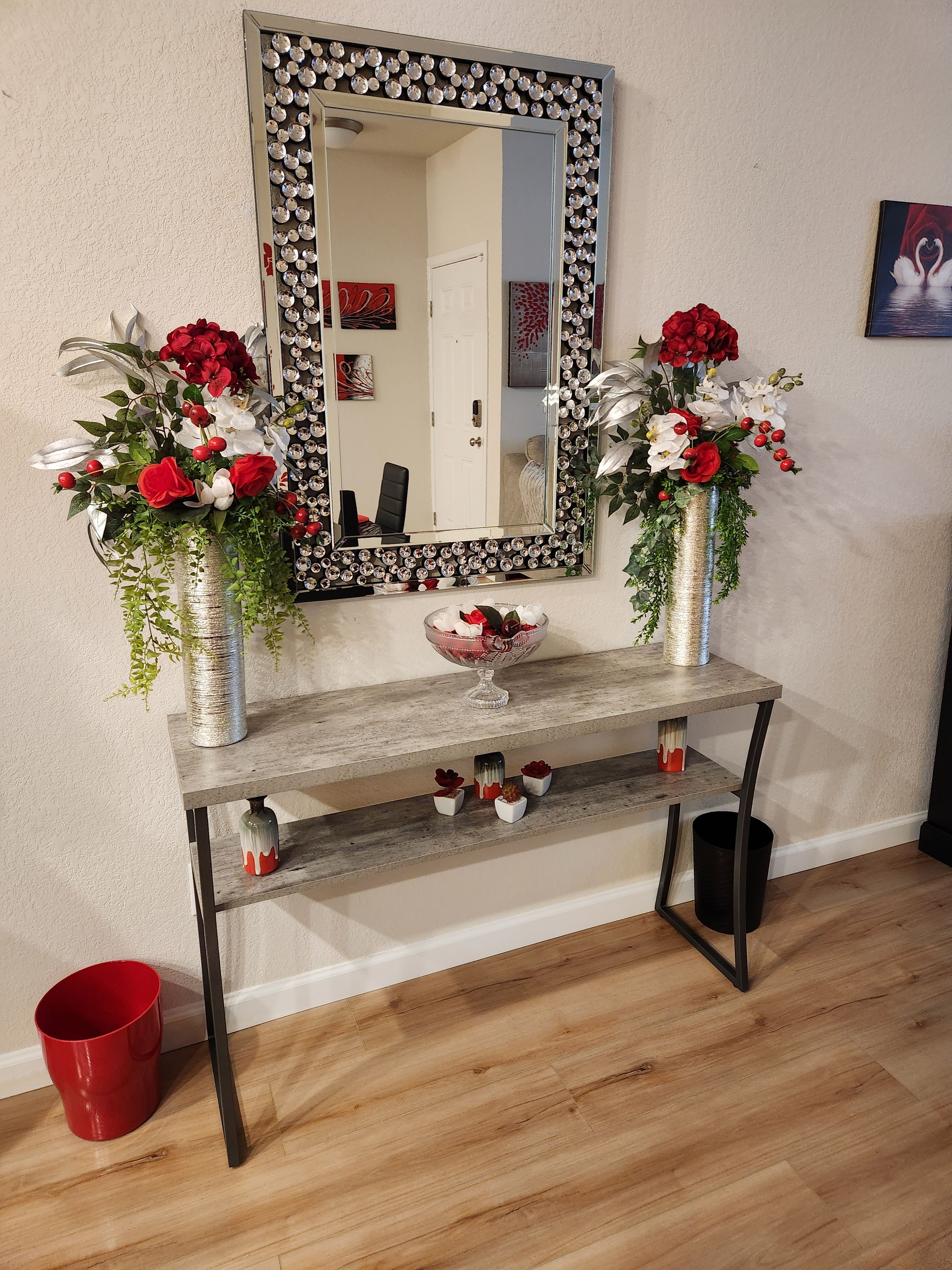 A console table with two vases of flowers, a mirror with a decorative frame, and a small shelf with decorative items.