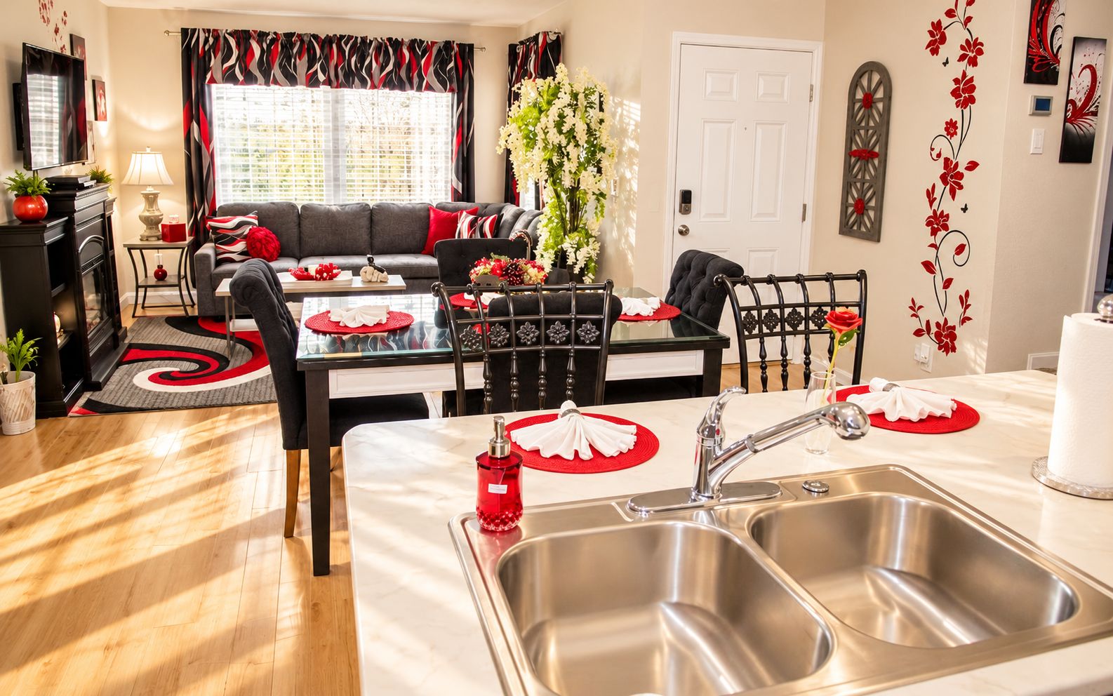 A modern kitchen with a stainless steel sink, a dining table with red placemats, and a black and red patterned window treatment.