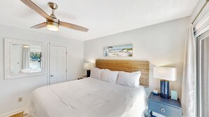 Ein Hotelzimmer mit einem Bett, einem Deckenventilator, einem Nachttisch mit Lampe, einem Gemälde und einem Fenster mit Vorhängen.
