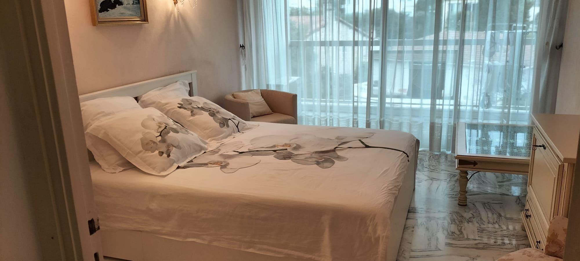 A bedroom with a bed, a nightstand, a chair, and a window with sheer curtains.