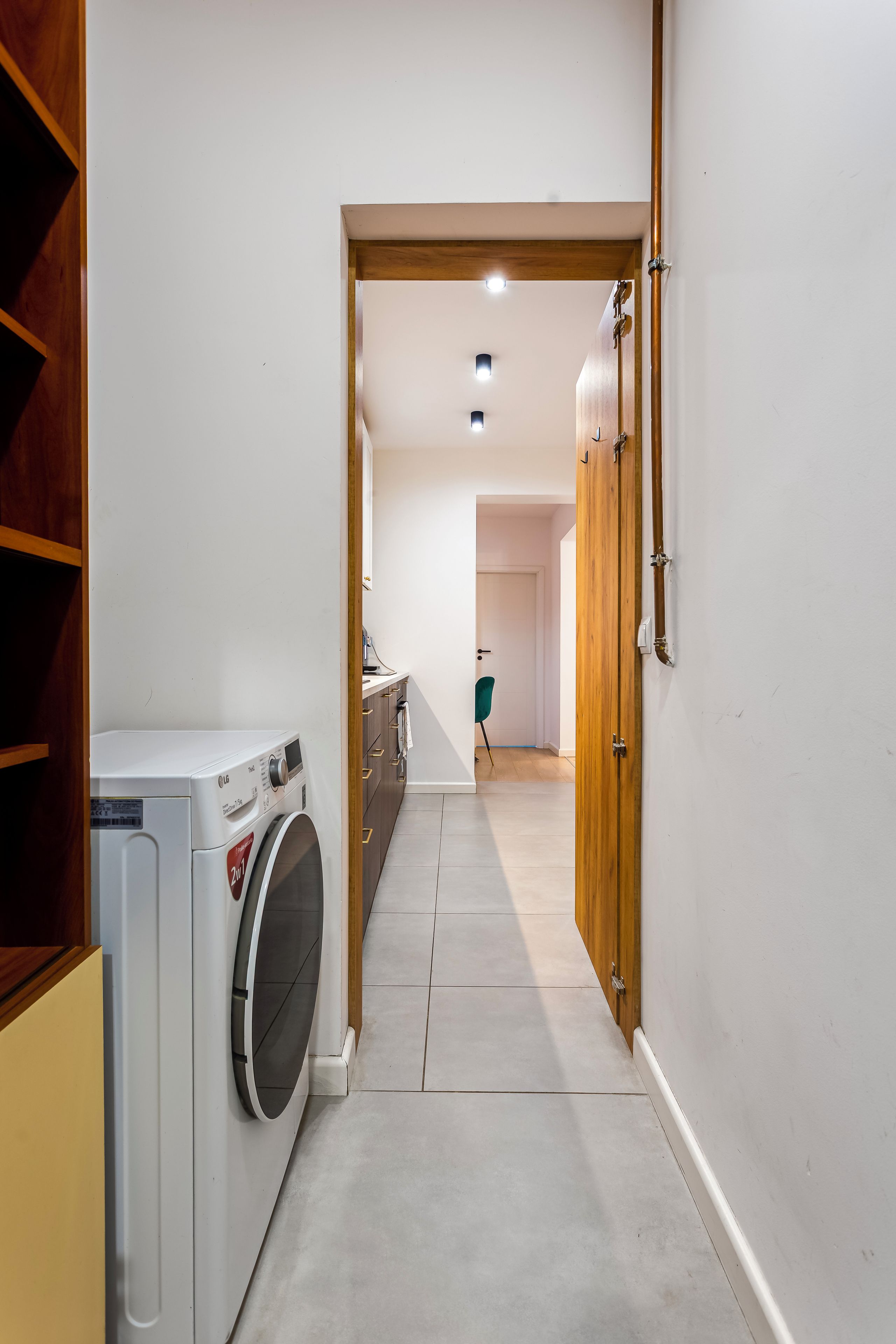Un pasillo estrecho con una lavadora a la izquierda, una puerta de madera a la derecha y una puerta que da a una habitación al final.