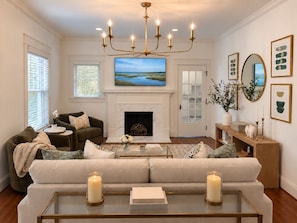 A living room with a fireplace, a large flat-screen TV, a chandelier, and a sofa. - Crimson Cottage | 1.4 Miles to Bryant-Denny! (Tuscaloosa)