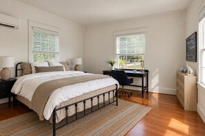 Ein Schlafzimmer mit Bett, Schreibtisch und Kommode, ausgestattet mit großen Fenstern und Blick ins Grüne.