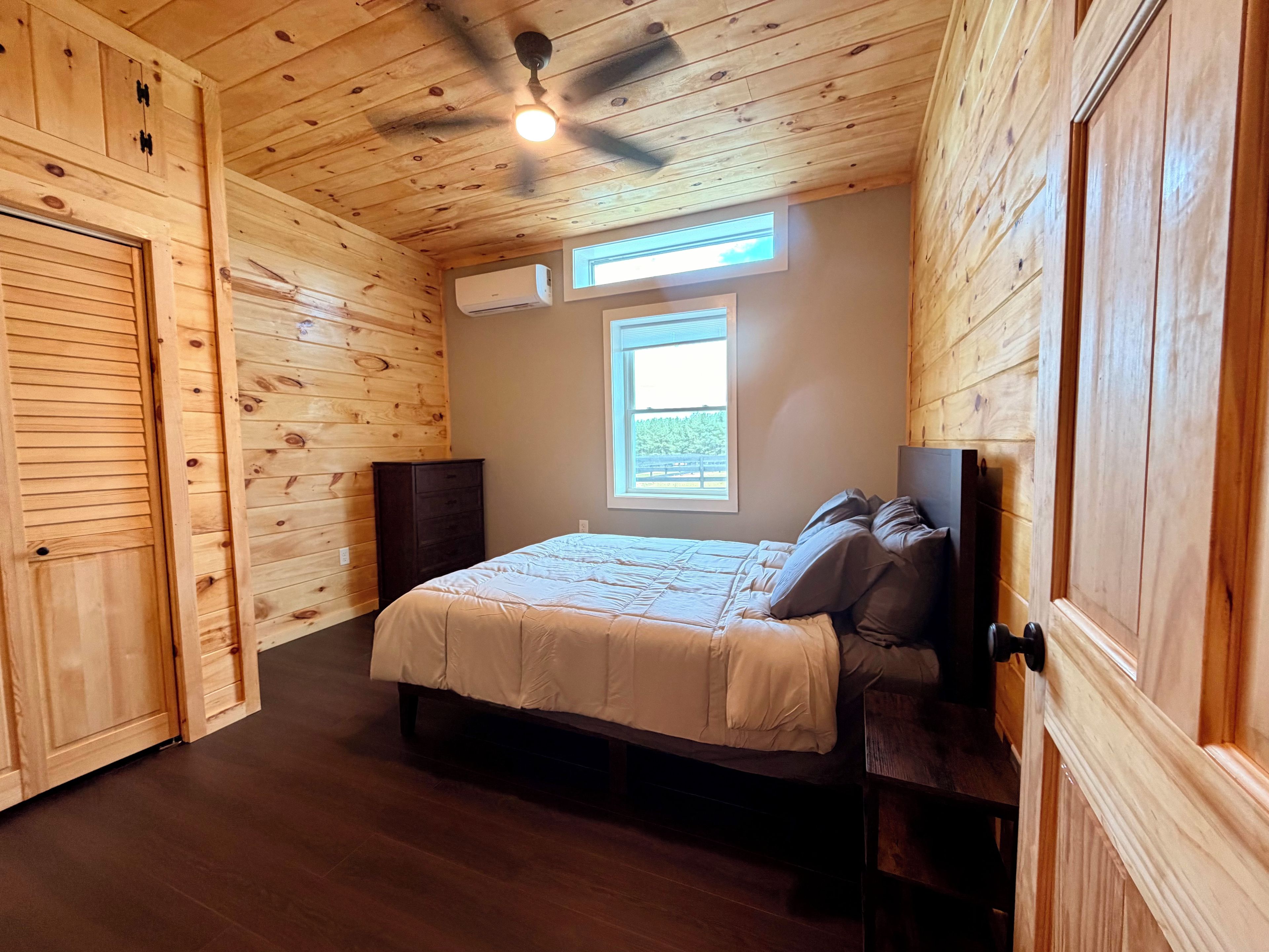 A bedroom with a wooden ceiling, a bed with a white bedspread, a window, and an air conditioning unit.