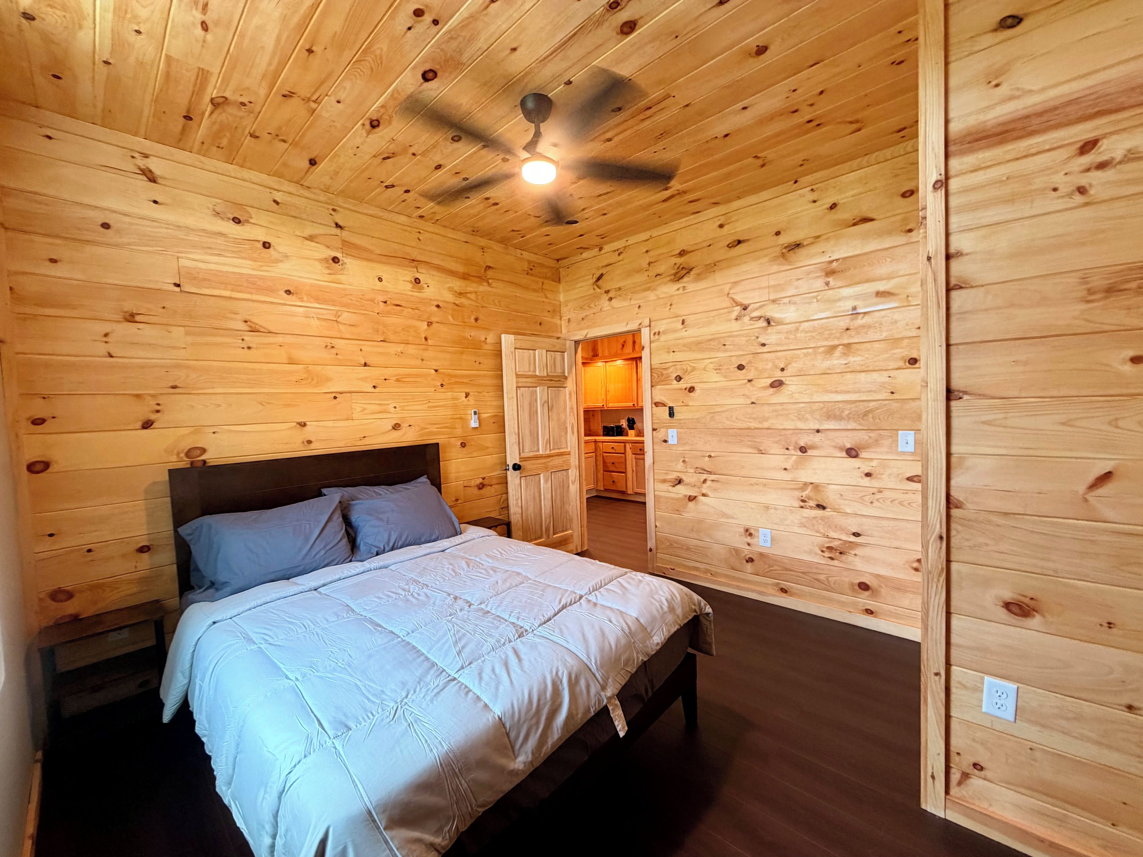 A wooden-paneled room with a bed, a ceiling fan, and a view into a kitchen area.