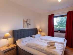 A hotel room with a large bed, two bedside tables with lamps, a window with red curtains, and a small plant on the windowsill. - Holiday Apartment With 1 Bedroom (Braunlage)