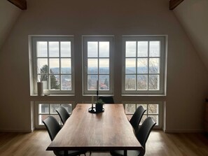 Ein Essbereich mit einem langen Holztisch und sechs Stühlen, drei Fenstern mit Blick auf eine Stadtlandschaft und einer Kerze auf dem Tisch.