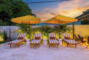 A patio with four striped lounge chairs under yellow umbrellas, surrounded by greenery and a white fence. - Palm Beach Retreat | Pool, Cold Plunge & Games (West Palm Beach)