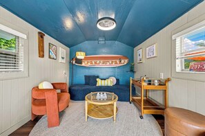 A compact living space with a blue ceiling, a surfboard decoration, a sofa, a coffee table, and a small wooden side table. - Palm Beach Retreat | Pool, Cold Plunge & Games (West Palm Beach)