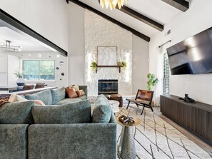 A modern living room with a sectional sofa, a fireplace, a television mounted on the wall, and a dining area in the background. - Jungle Oasis - Pool + Hot Tub + Game Rooms (Richardson)