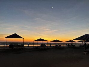 Una playa con varios sombrillas y sillas de playa, cielo despejado y una luna creciente.