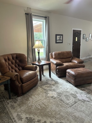 A living room with a brown leather sofa, a matching ottoman, a small table with a lamp, and a patterned rug.
