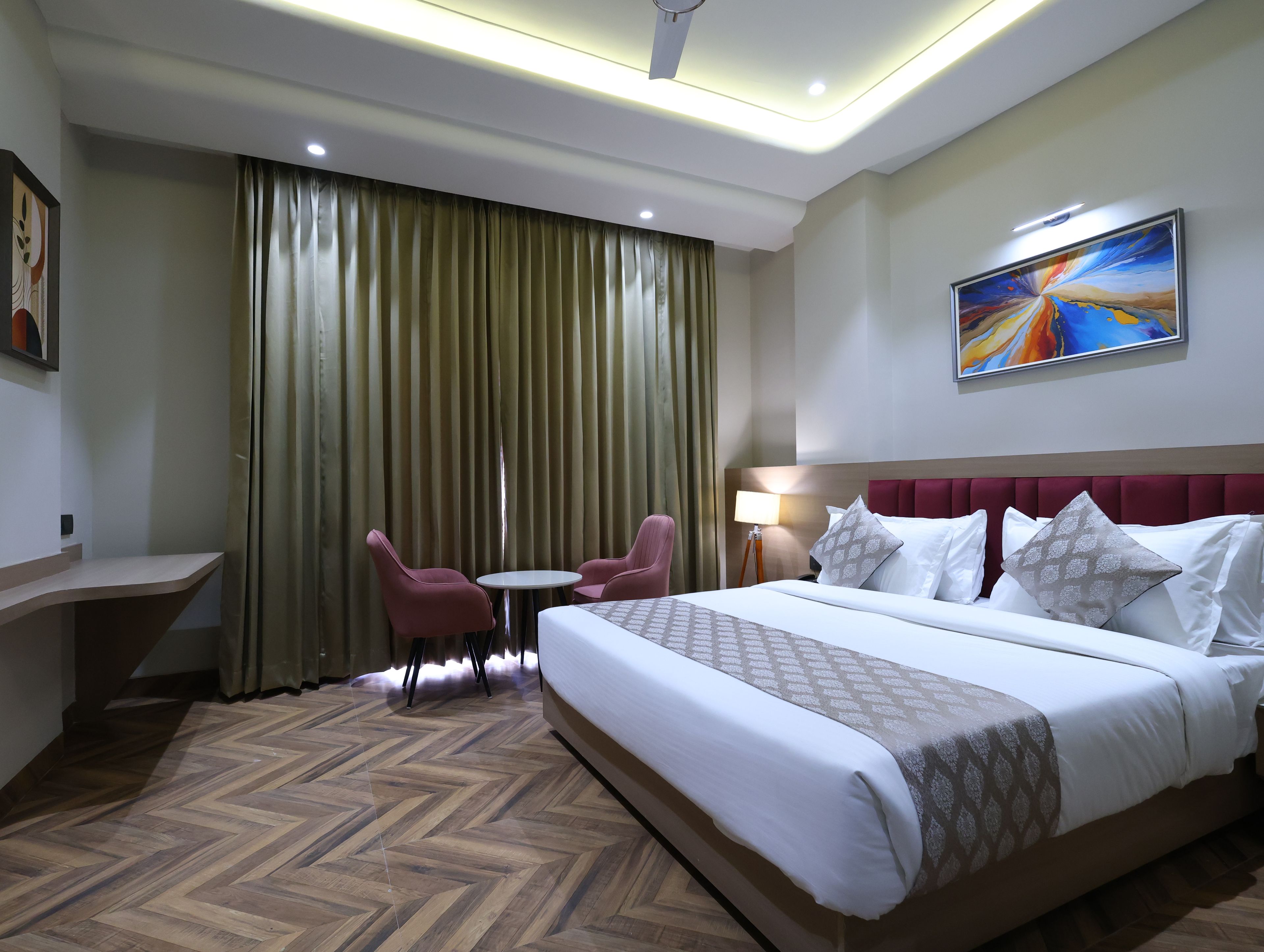 A hotel room with a large bed, two chairs, a small table, a desk, a lamp, and a painting on the wall.