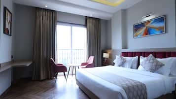A hotel room with a large bed, a desk, a chair, a window with curtains, a painting on the wall, and a ceiling fan.