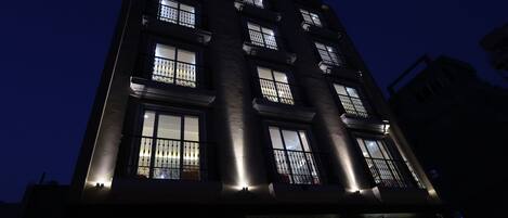 Een meerhoog gebouw met verlichte ramen en balkons, een bord met 'The Kailash Premium Hotel' en een zichtbare ingang met een glazen deur.