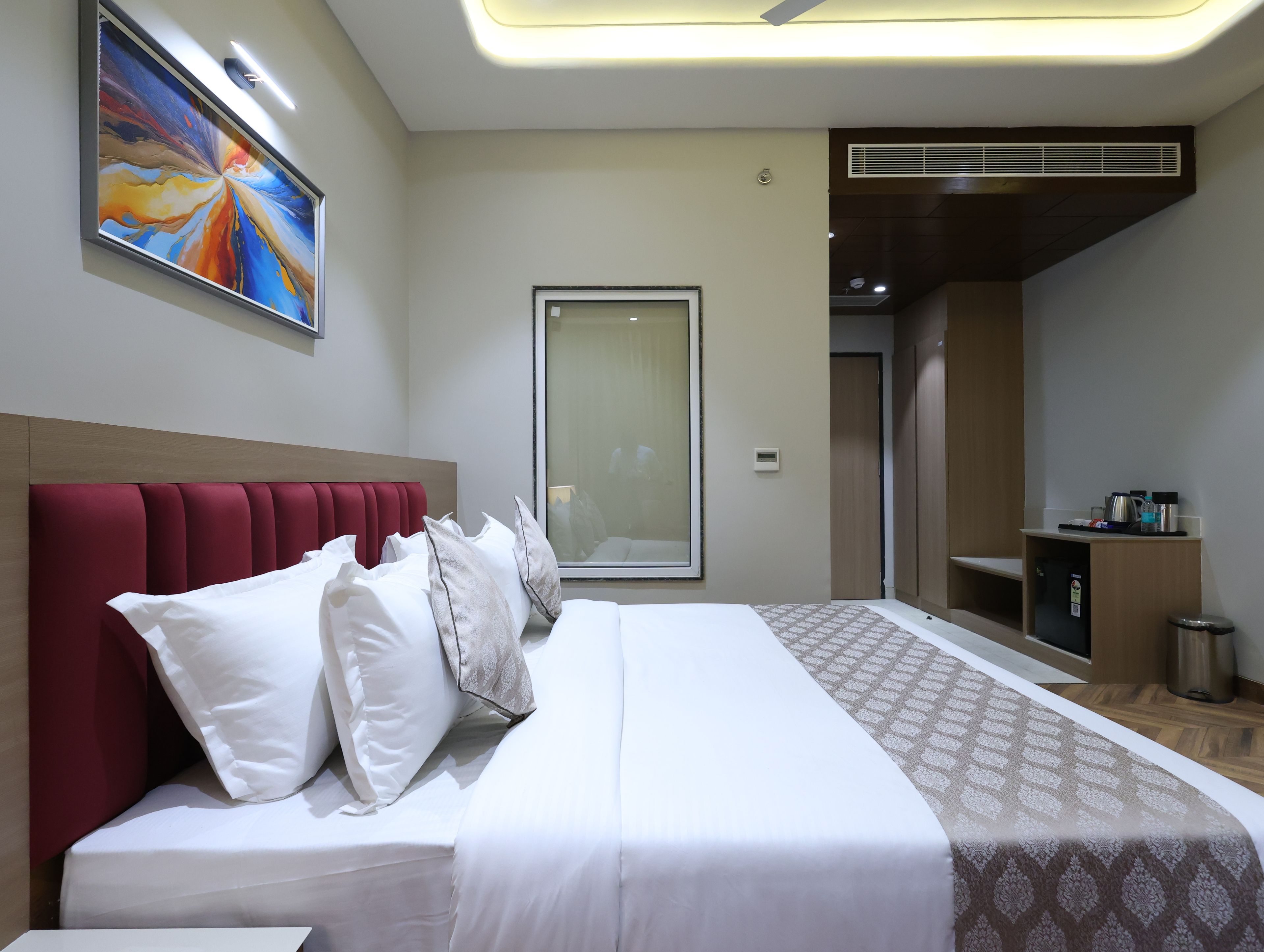 A hotel room with a bed, a large mirror, a painting on the wall, and a kitchenette area.