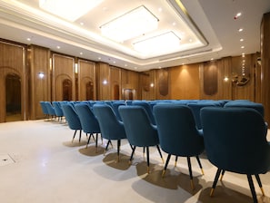 Un salón de conferencias con filas de sillas azules, paneles de madera e iluminación empotrada en el techo.