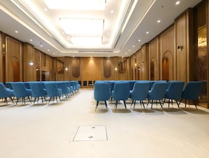 Sala de conferências com fileiras de cadeiras azuis, paredes de painel de madeira e iluminação embutida no teto.
