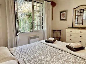 A bedroom with a bed, a dresser, a chair, a radiator, and a window with curtains. - Holiday Home With 1 Bedroom (Entrecasteaux)
