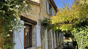 A stone building with a window and shutters, surrounded by greenery and a gravel path.