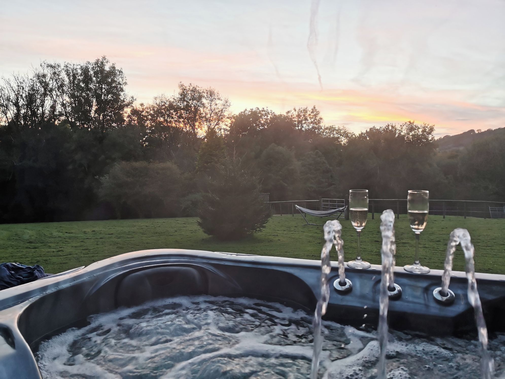 A hot tub with water spouting from its nozzles, surrounded by a grassy area with trees and a fence in the background.