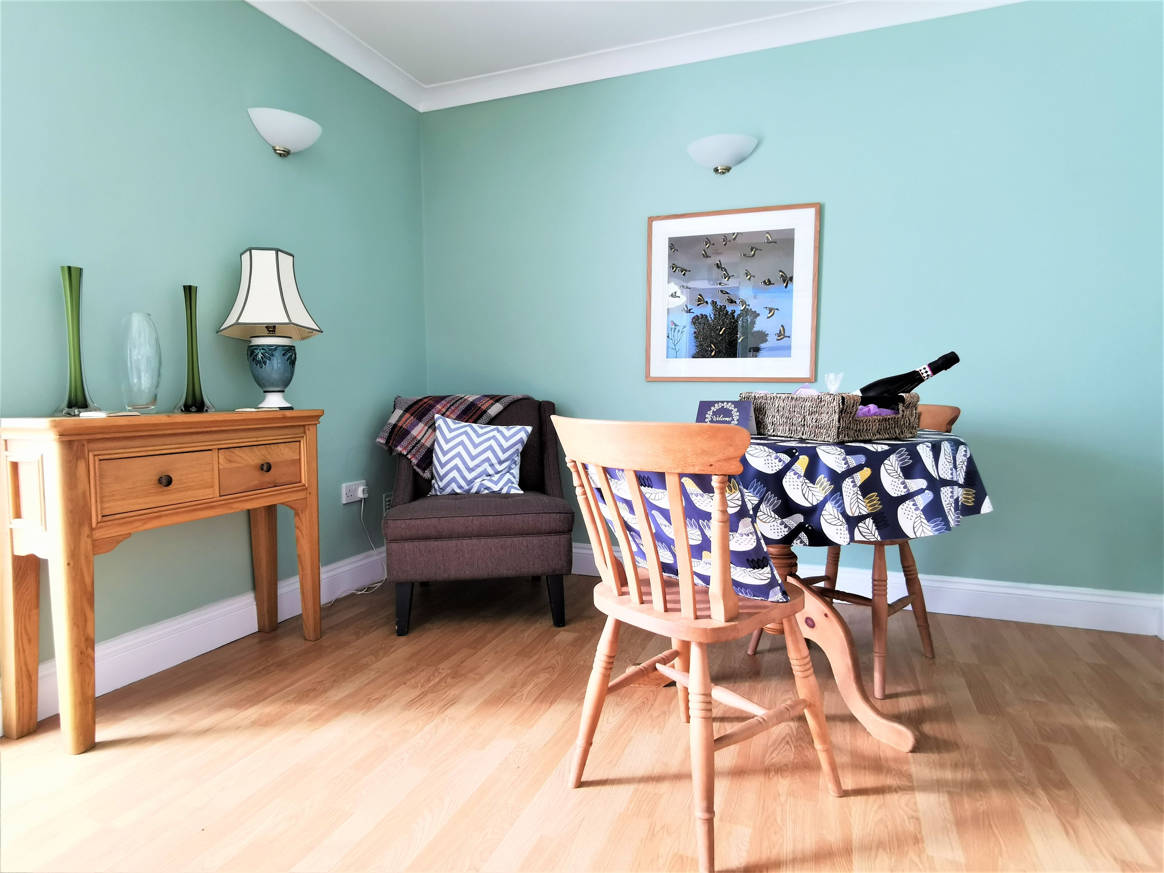 Una habitación con paredes verde azulado, piso de madera, una mesa de comedor con mantel estampado, una silla, una lámpara y un cuadro en la pared.