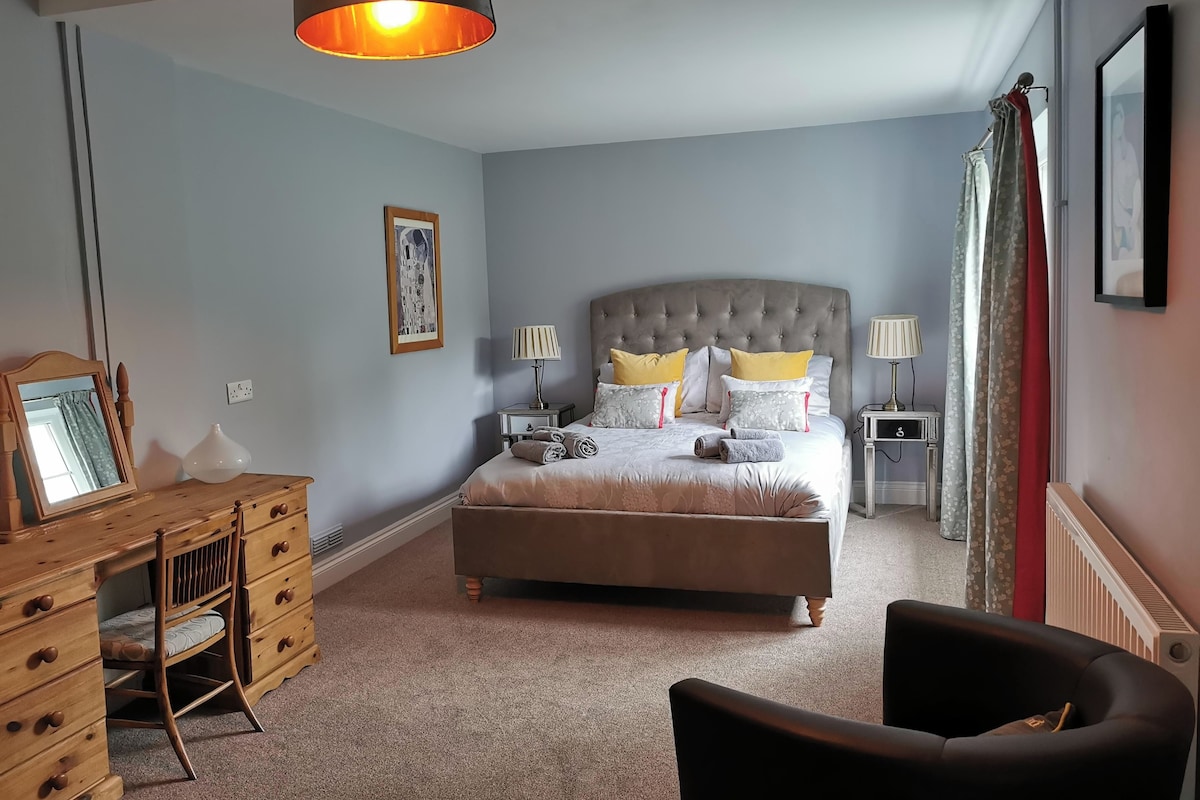 Ein Schlafzimmer mit einem Bett, Nachttischen, einem Schreibtisch, einem Sessel und einer Pendelleuchte.