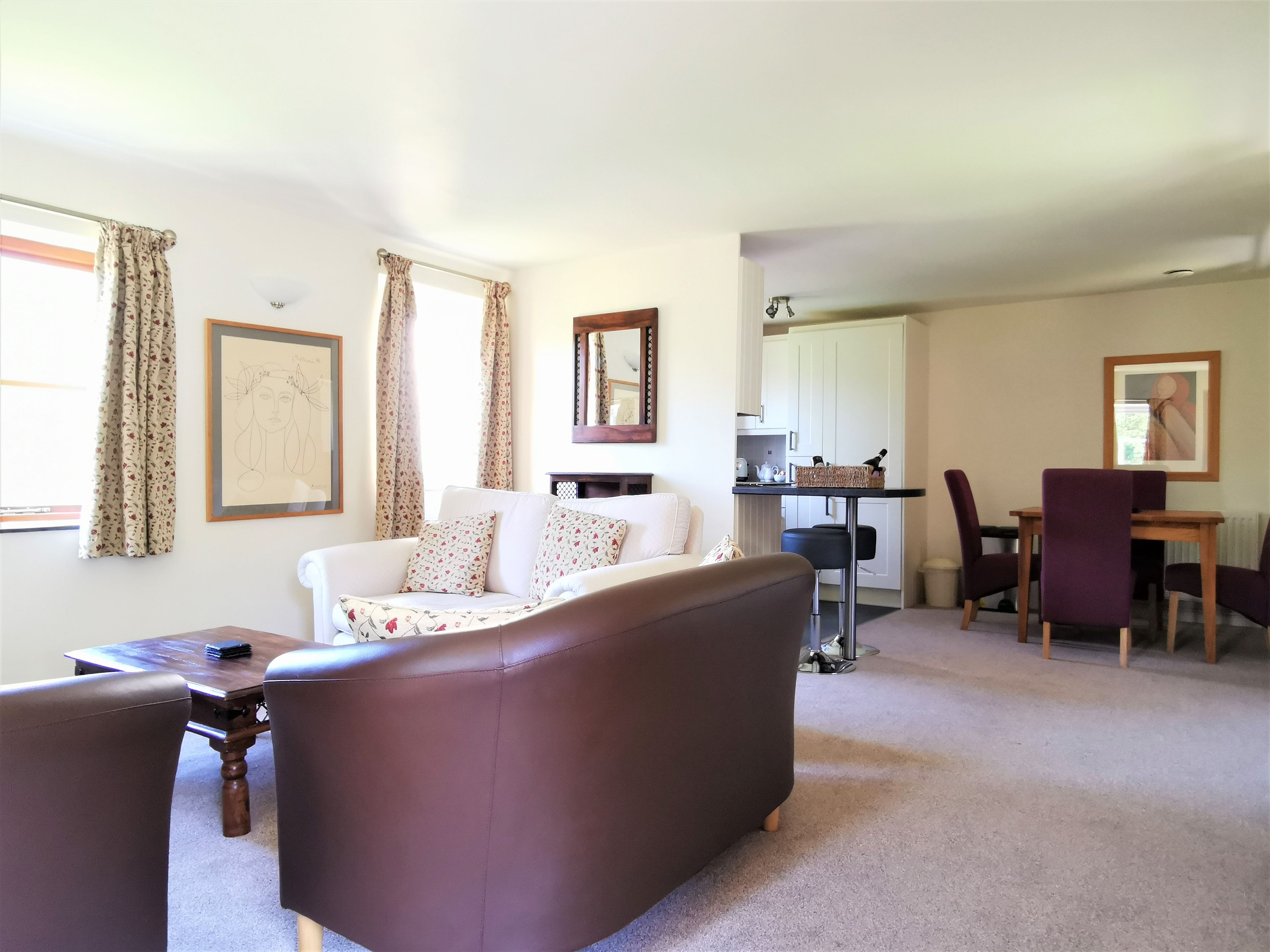 A living room with a brown sofa, a white sofa, and a dining table with purple chairs. There is a kitchen area with a white cabinet and a hanging light fixture.