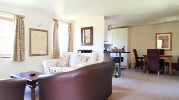 A living room with a brown sofa, a white sofa, and a dining table with purple chairs. There is a kitchen area with a white cabinet and a hanging light fixture.