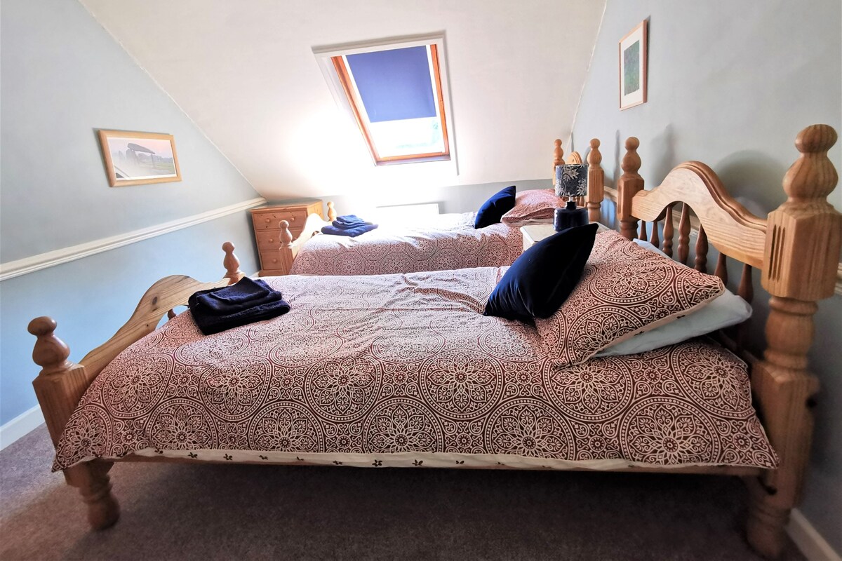 Ein Schlafzimmer mit zwei Holzbetten, gemusterten Betttextilien, einem kleinen Fenster und einem Nachttisch.