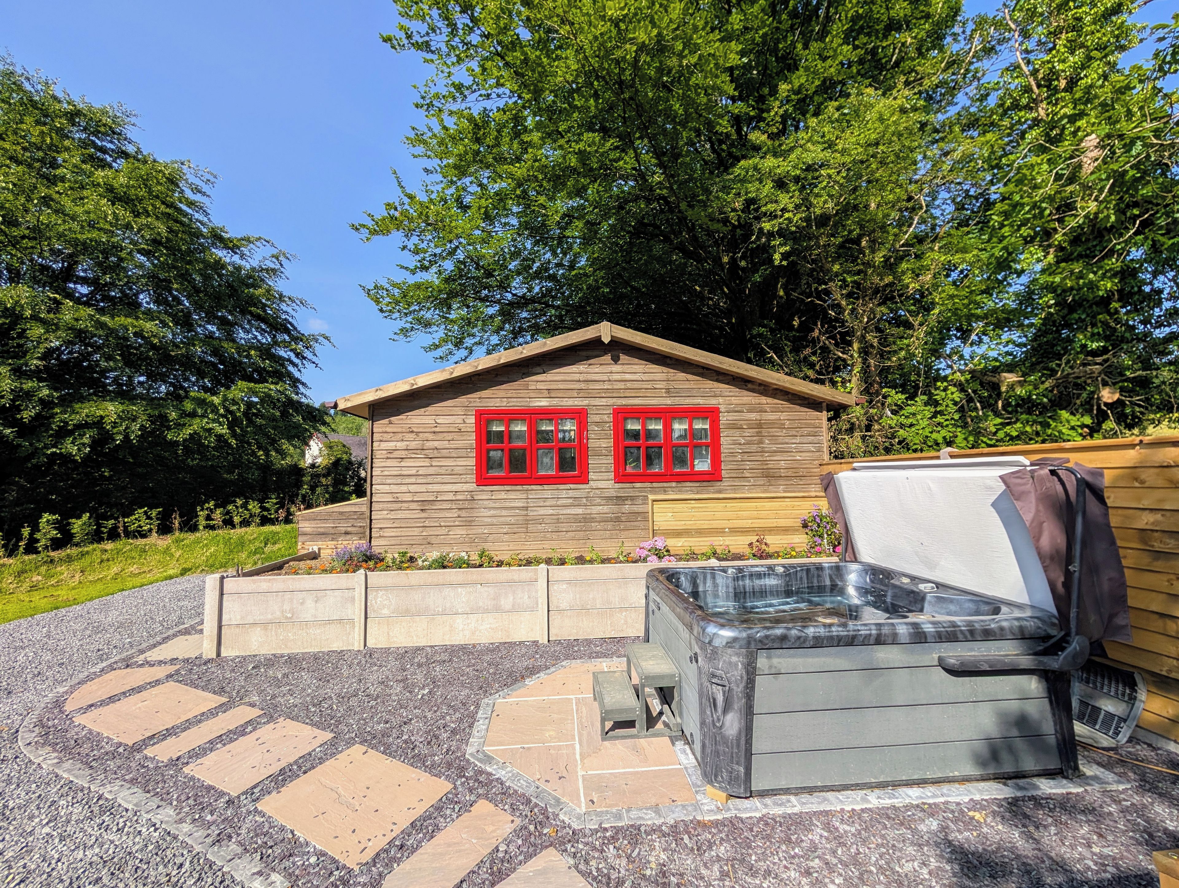A wooden cabin with red-framed windows, a hot tub, and a gravel pathway.
