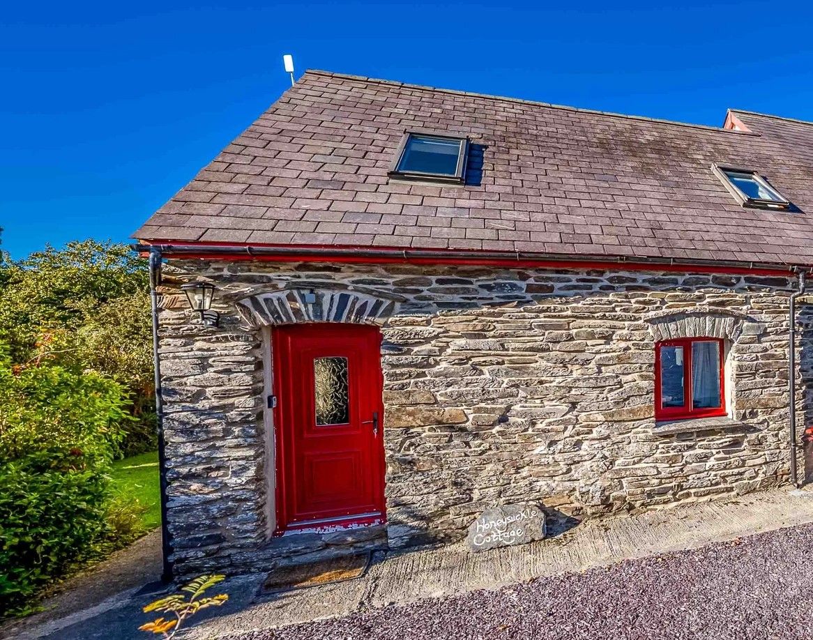 Una cabaña de piedra con puerta roja y dos ventanas, rodeada de follaje.