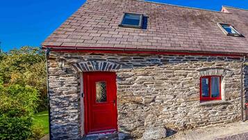 Una cabaña de piedra con puerta roja y dos ventanas, rodeada de follaje.