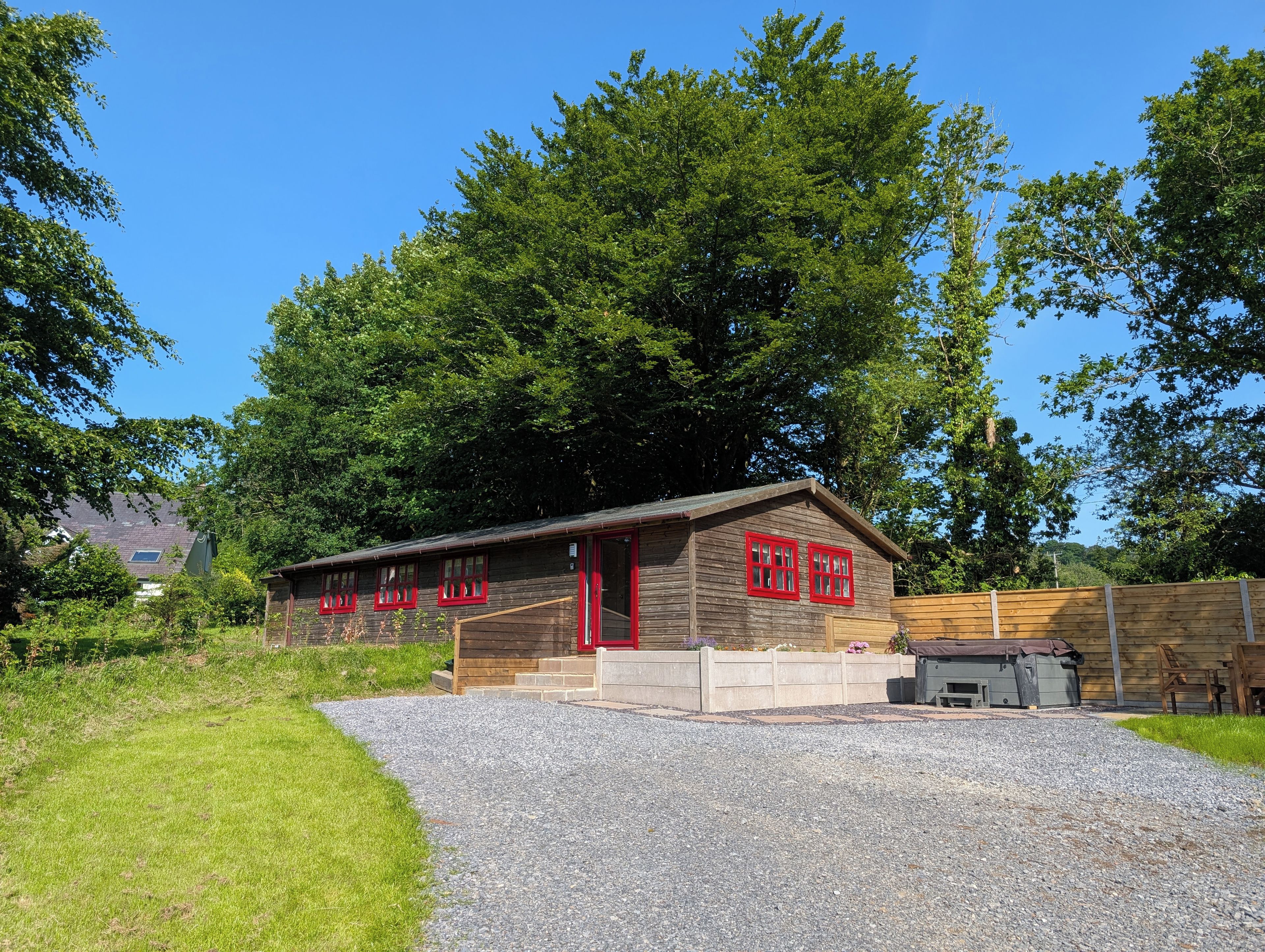A wooden cabin with a gravel driveway, surrounded by trees and a clear sky.