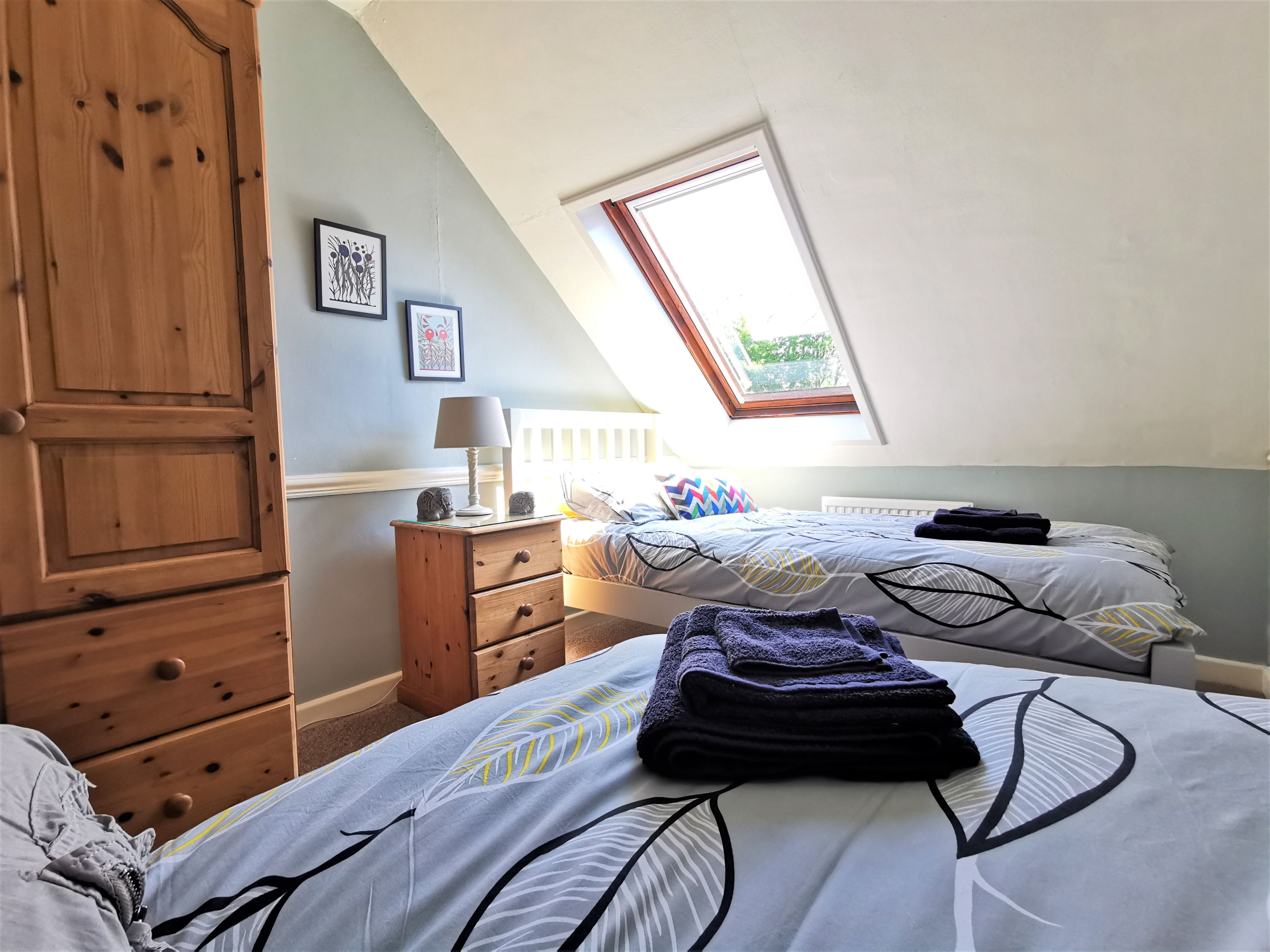 A bedroom with two beds, a wooden wardrobe, a nightstand, and a window with a view of greenery.