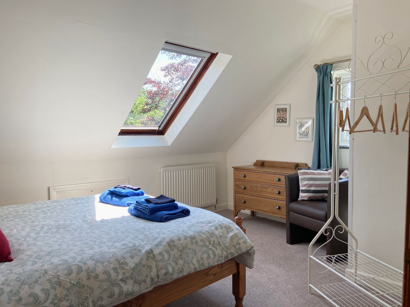 Un dormitorio con tragaluz, cama con toallas dobladas, cómoda, silla y un tendedero de metal.