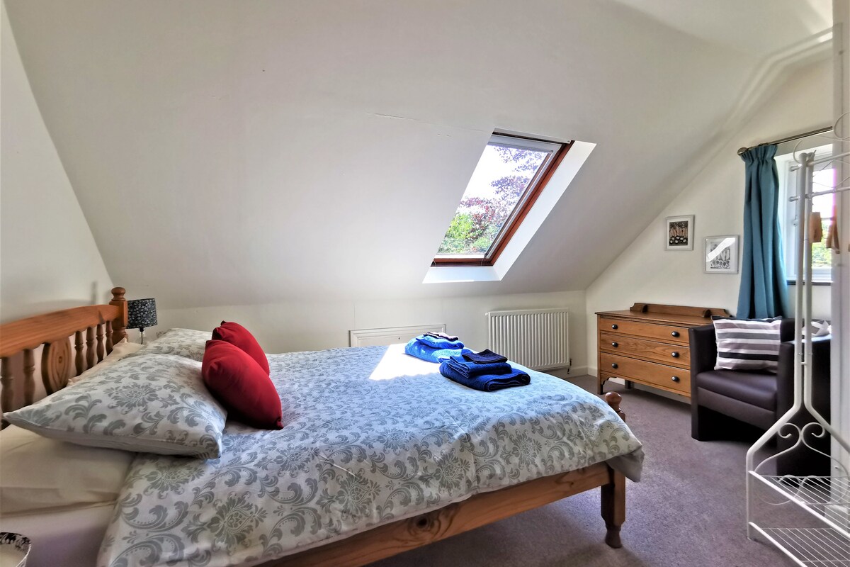 Ein Schlafzimmer mit schrägen Wänden, einem Oberlicht, einem Bett mit gemusterten Betttextilien, einem Schminktisch und einem Stuhl.
