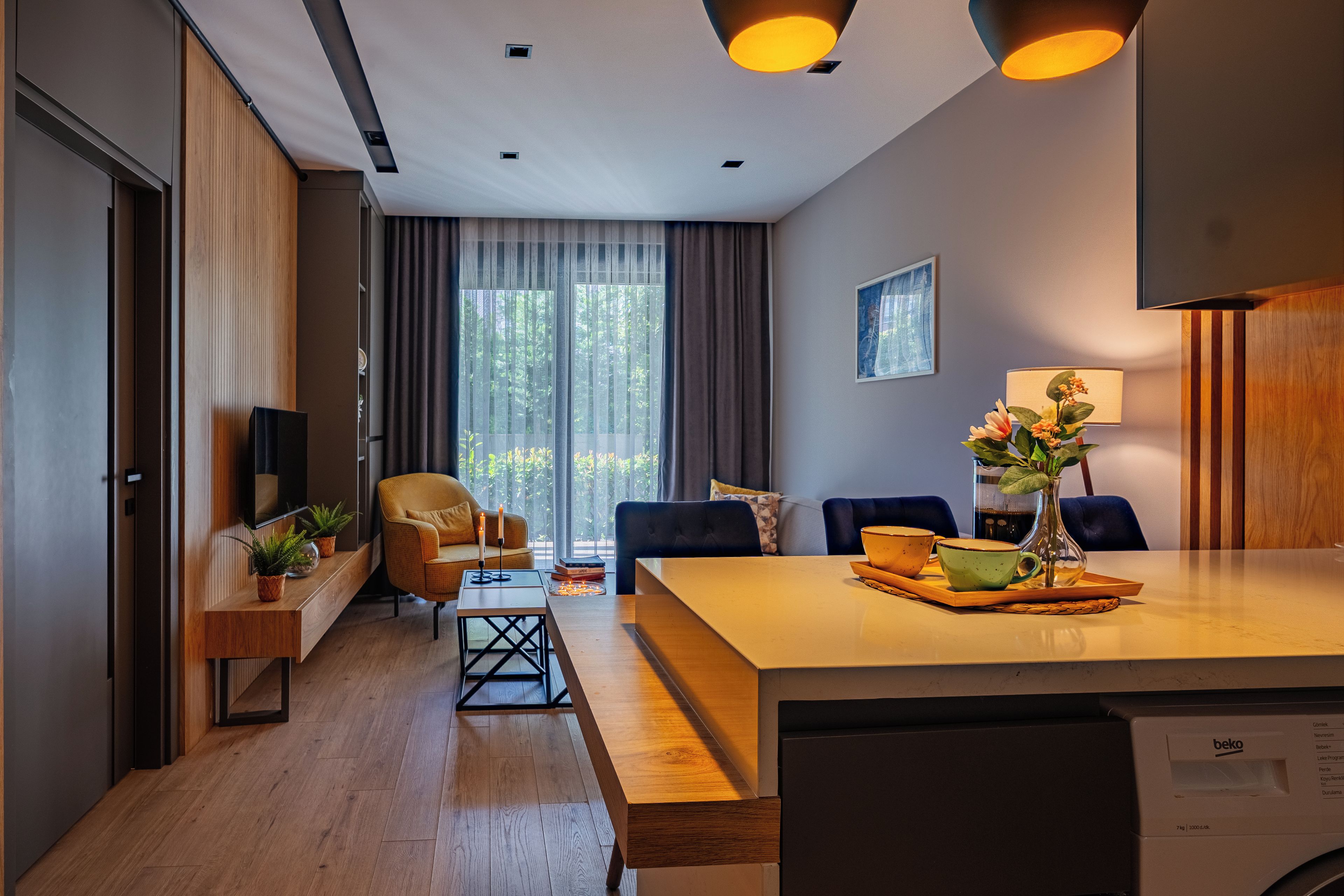 A modern hotel room with a wooden floor, a dining area with a table and chairs, a sofa, a TV, and a window with curtains.