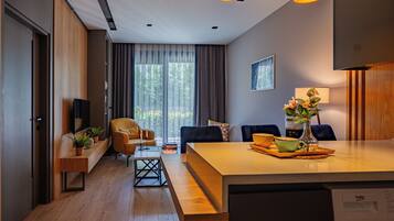 A modern hotel room with a wooden floor, a dining area with a table and chairs, a sofa, a TV, and a window with curtains.