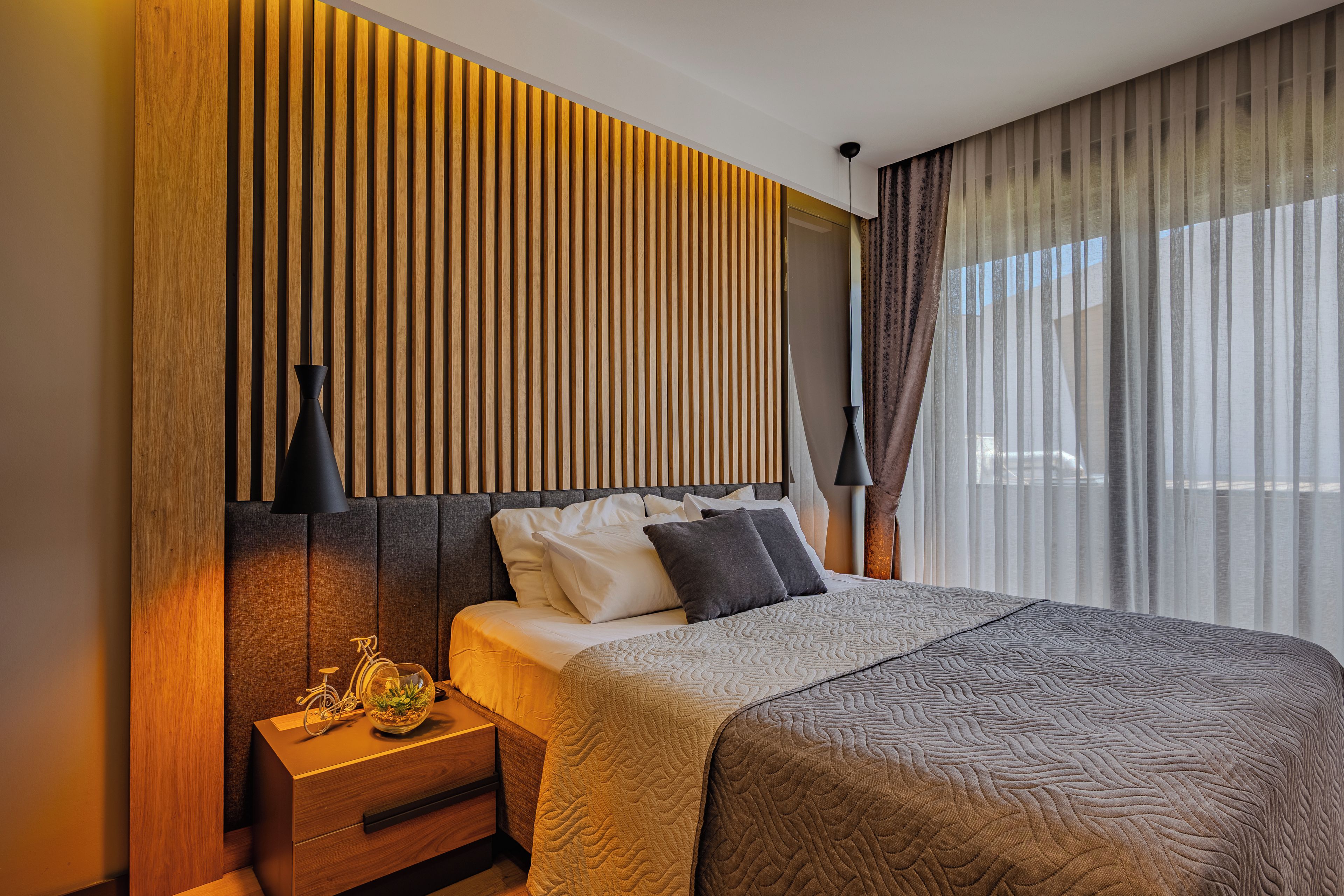Una camera d'albergo moderna con parete in doghe di legno, un letto con copripiumino trapuntato, un comodino con una lampada e una finestra con tende leggere.