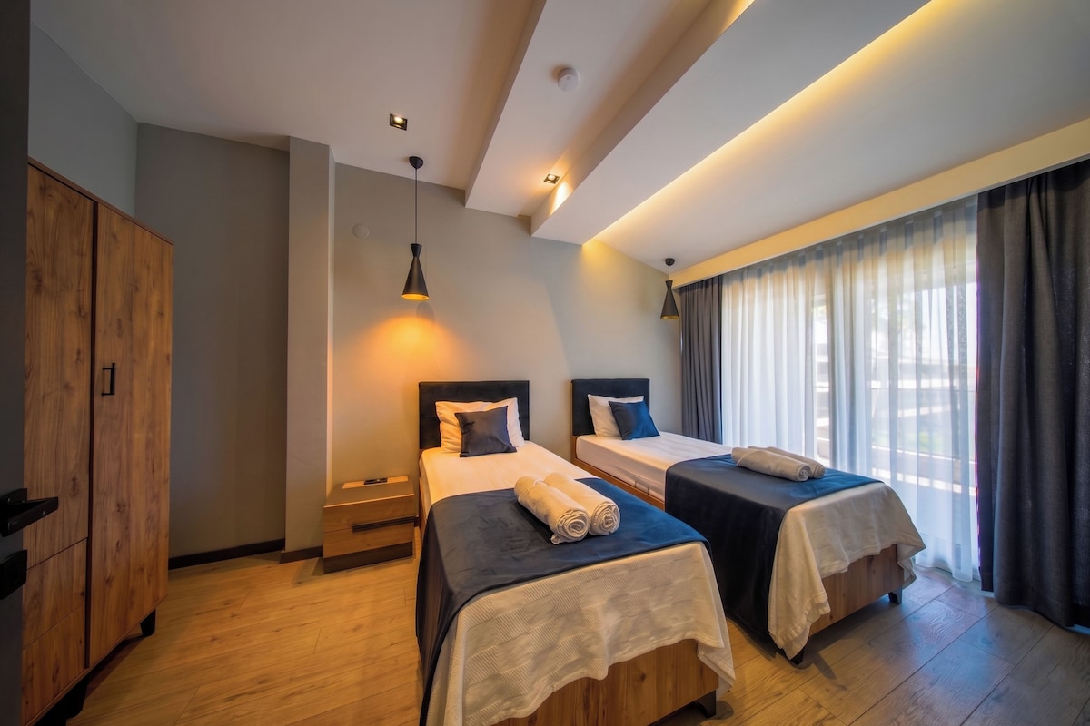 A hotel room with two beds, wooden flooring, a wardrobe, and a window with curtains.
