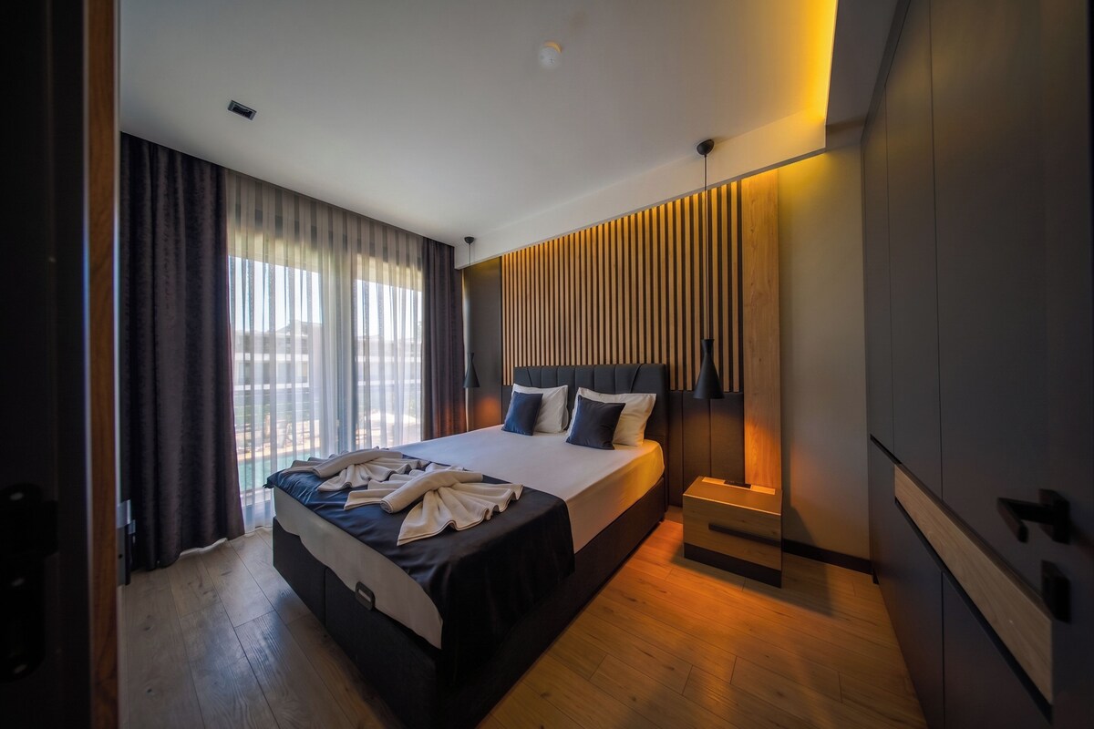 A modern bedroom with a large bed, a bedside table, and a view of the outdoors through the window.